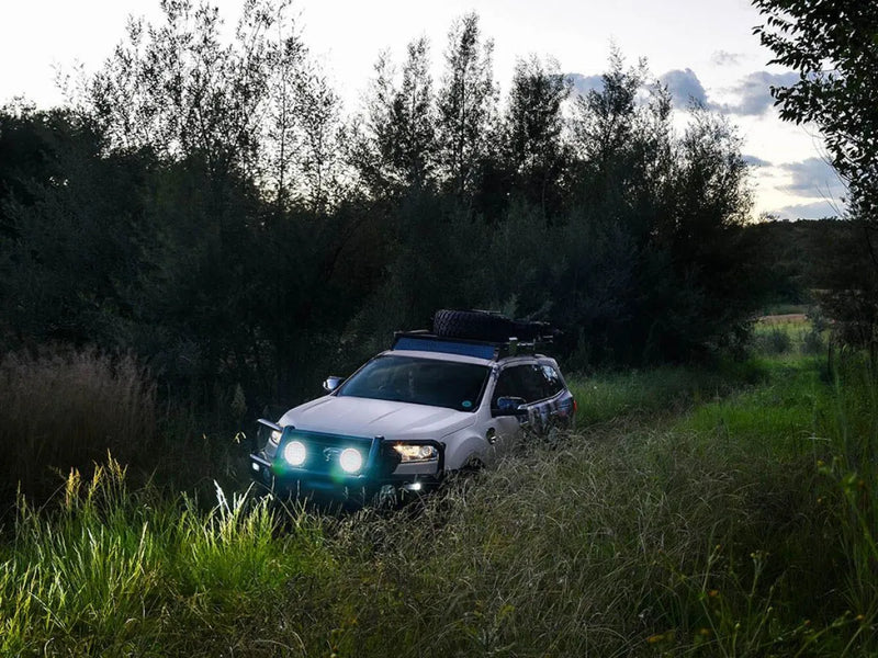 Silver SUV with bright headlights in tall grass showcasing Front Runner Ford Everest Slimline II Roof Rack Kit