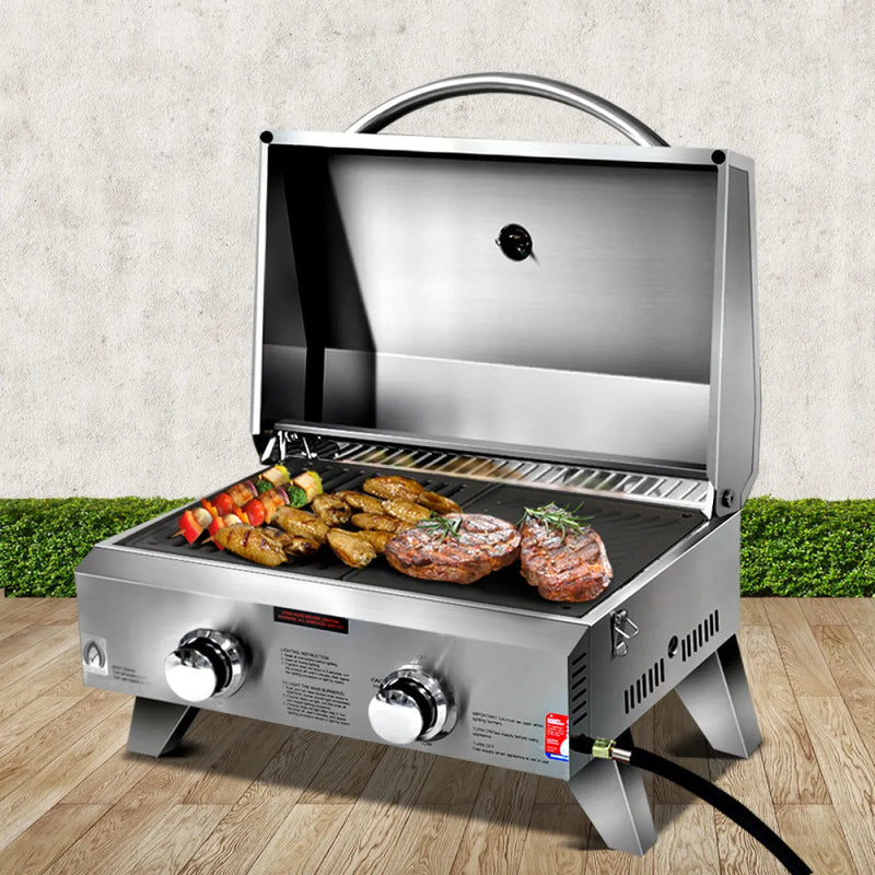 Portable stainless steel gas bbq with open lid showing grilled meats and vegetables